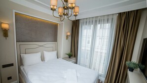 A neatly made bed with white linens, a modern chandelier, and a large window with curtains.