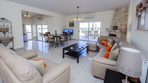 Ein geräumiges Wohnzimmer mit einer Couch, einem Couchtisch und einem Essbereich. Es gibt einen Fernseher, einen Kamin und einen Ausblick ins Freie.