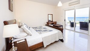 Ein Schlafzimmer mit Bett, Nachttisch, Lampe, Kommode und Blick auf das Meer durch eine Schiebetür.