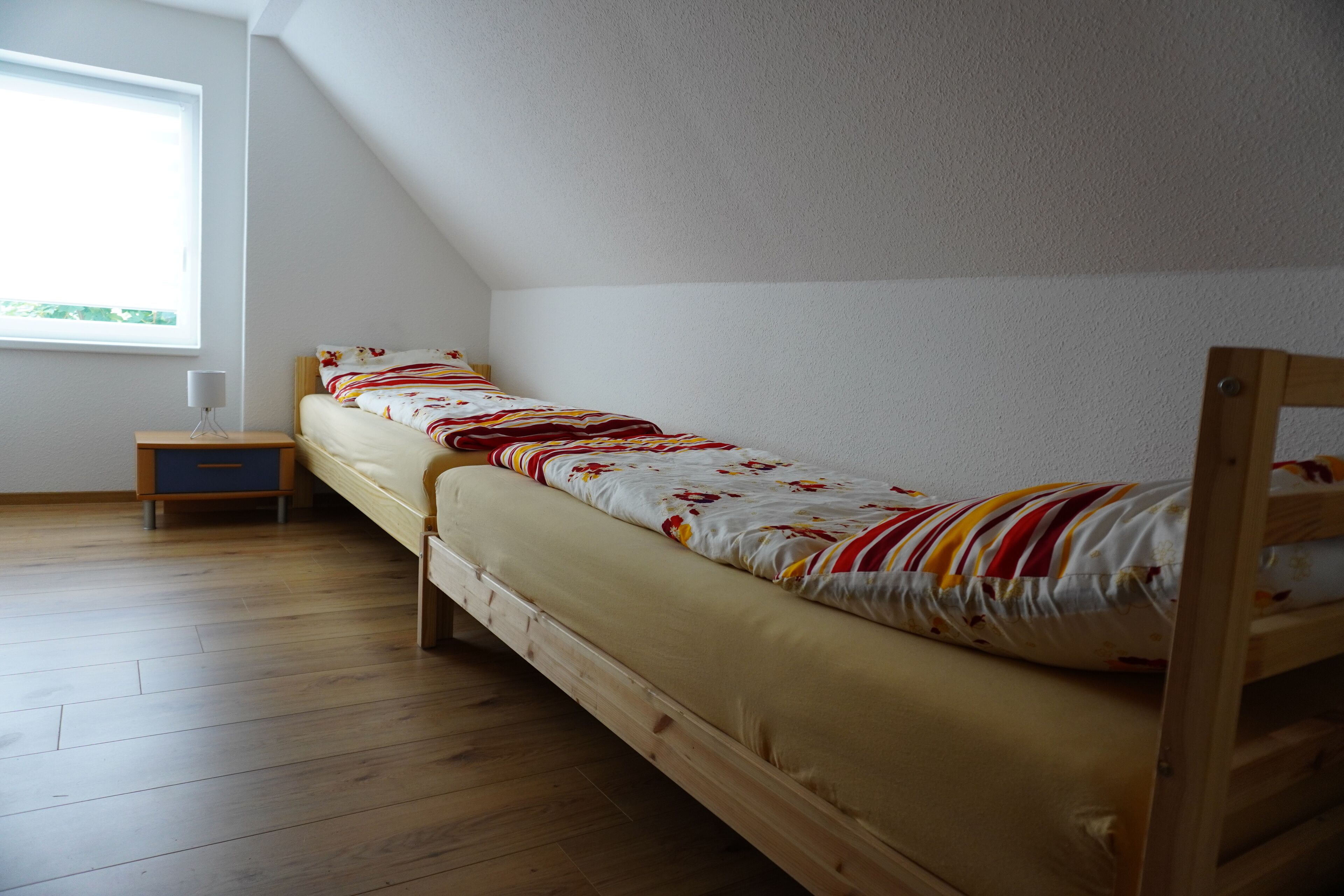 Ein Einzelbett mit Holzrahmen, ein kleiner Nachttisch und ein Fenster mit Tageslicht.