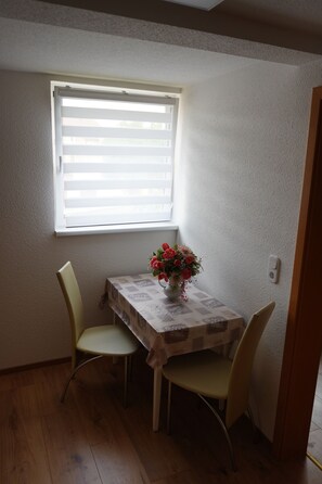 A small dining area with a table and two chairs, a window with blinds, and a vase with flowers on the table.