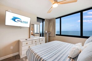 A hotel room with a bed, a dresser, a mirror, a ceiling fan, and a large window offering a view of the ocean.