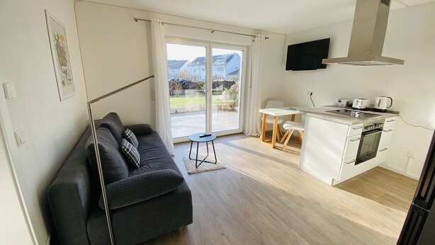 Un apartament modern amb un sofà negre, una taula de dinar petita, una cuina amb fogó i microones, i una gran porta corredissa de vidre que dona a un balcó.
