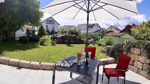 Un pati amb una taula i cadires, un parasol blanc, i vista a un jardí amb cases al fons.