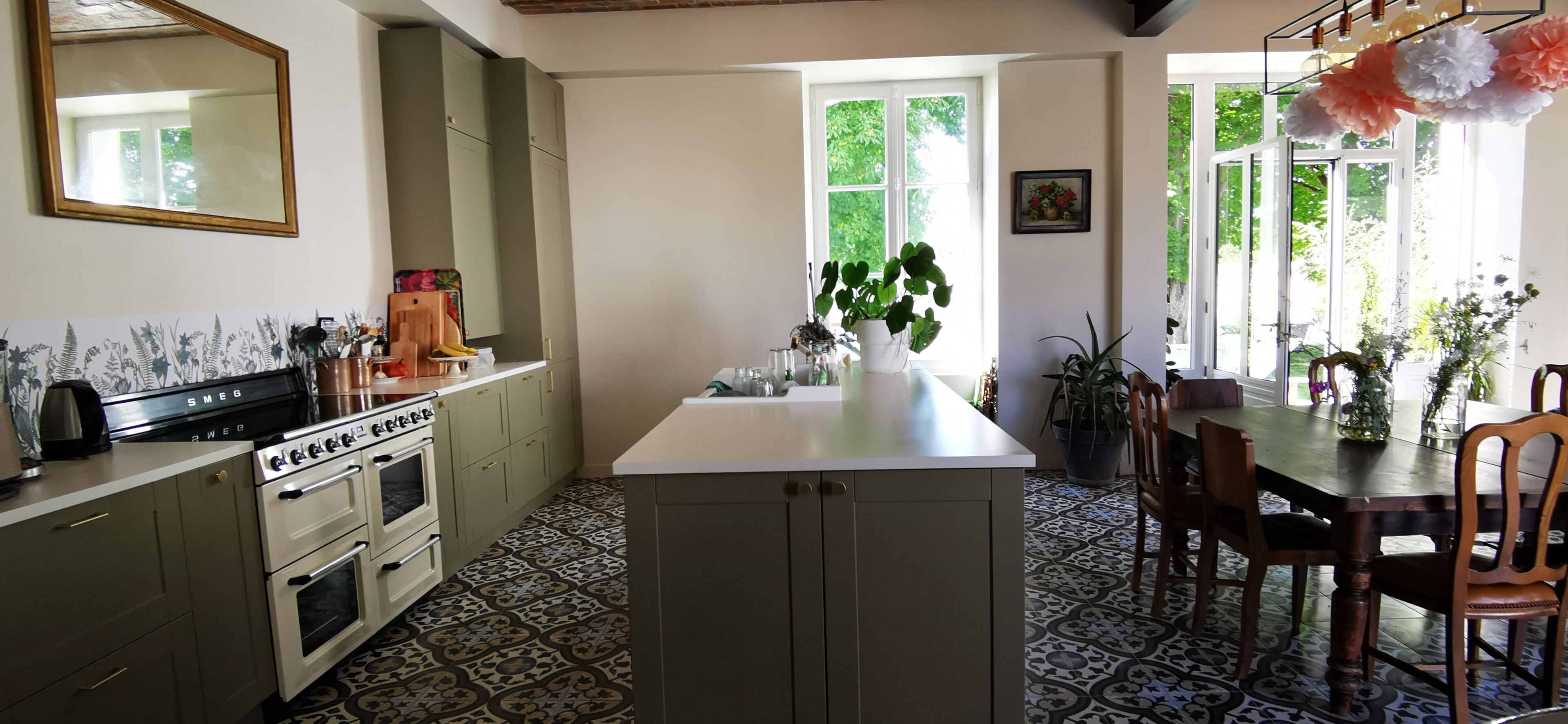 Een keuken met een centraal eiland, een eethoek met houten stoelen en een groot raam met uitzicht op groen.