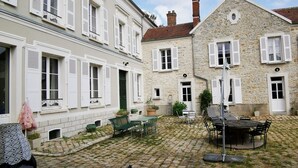 Une cour dallée de pierres, avec des sièges extérieurs et des bâtiments en pierre dotés de volets blancs.