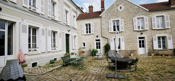 A courtyard with cobblestone flooring, outdoor seating, and stone buildings with white shutters.