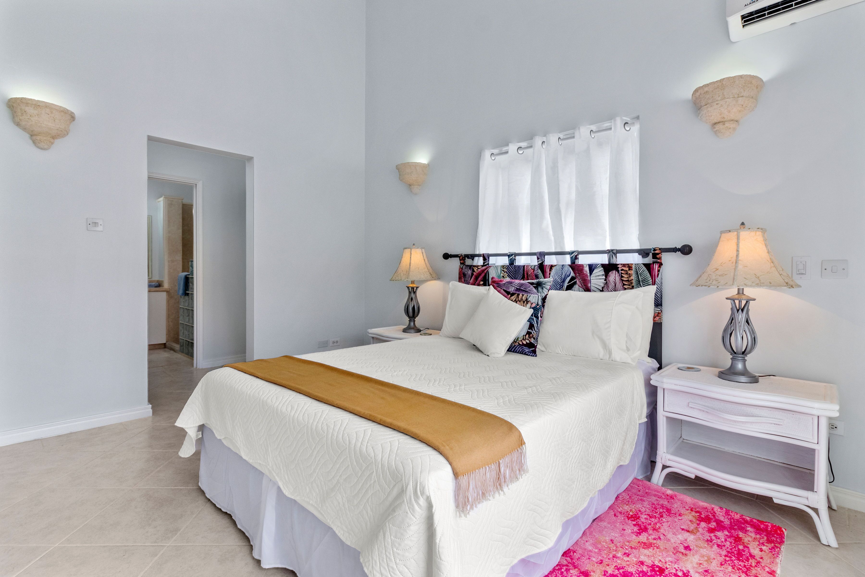 Un dormitorio con una cama, dos mesitas de noche con lámparas, una cortina blanca y una puerta que da a otra habitación.