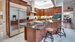 A modern kitchen with wooden cabinets, a stainless steel refrigerator, a marble island, and bar stools. - Golf Cart, Bimini Cove by SeaBreeze Vacation (Anna Maria)