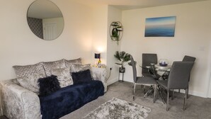 A living room with a grey couch, a blue throw blanket, a round mirror, a dining table with chairs, a small plant, and a painting on the wall. - Woodside View (Whitby)