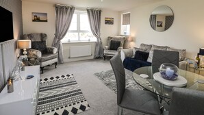 A cozy living room with a fireplace, comfortable seating, a dining area, and a large window with curtains. - Woodside View (Whitby)