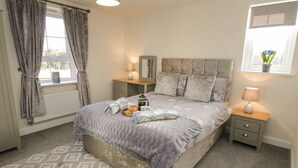 A bedroom with a bed, nightstands, a window with curtains, a radiator, and a patterned carpet. - Woodside View (Whitby)