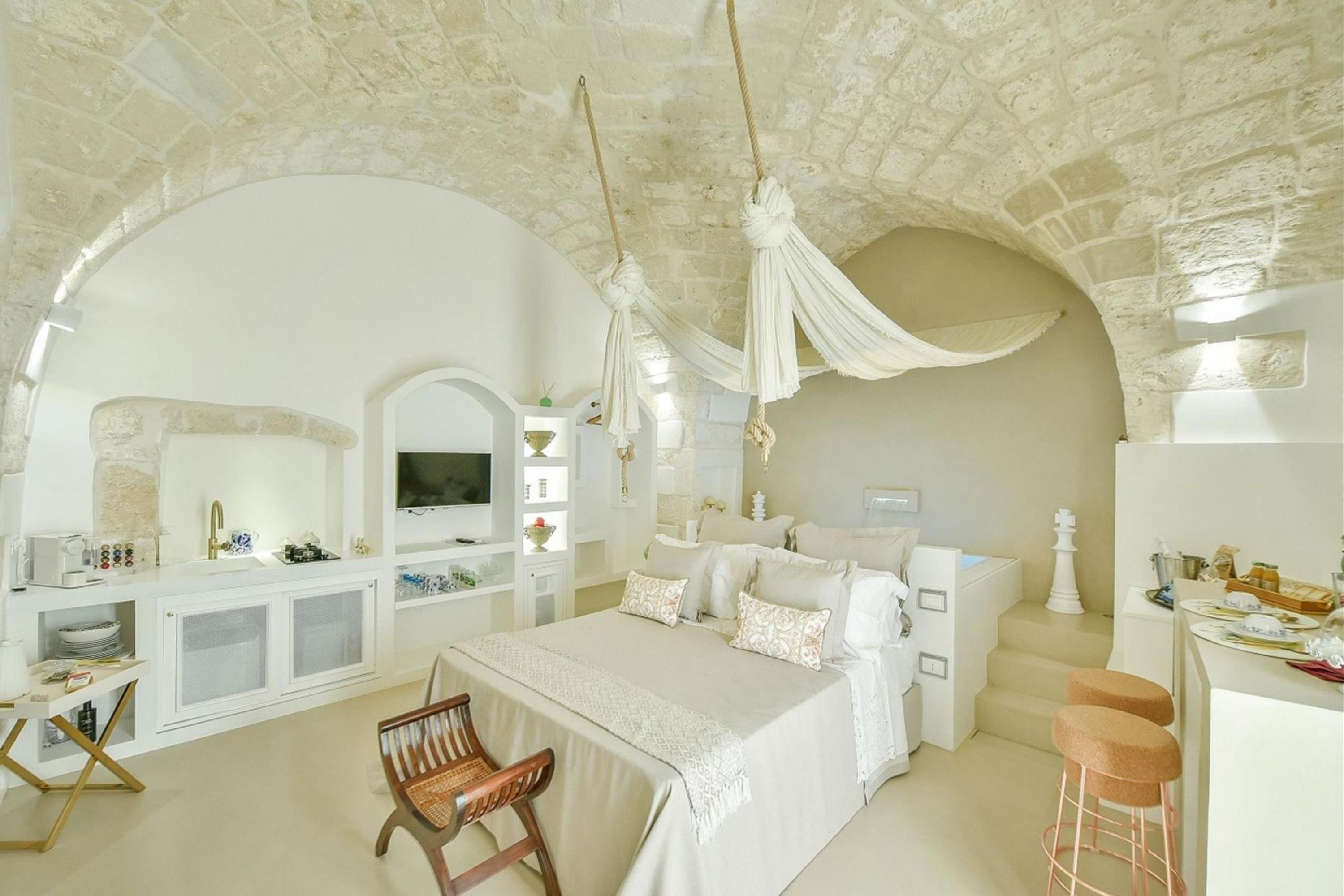 Un'ampia camera da letto con un grande letto, una scrivania e una sedia. Il soffitto è in pietra e c'è una TV appesa al muro.