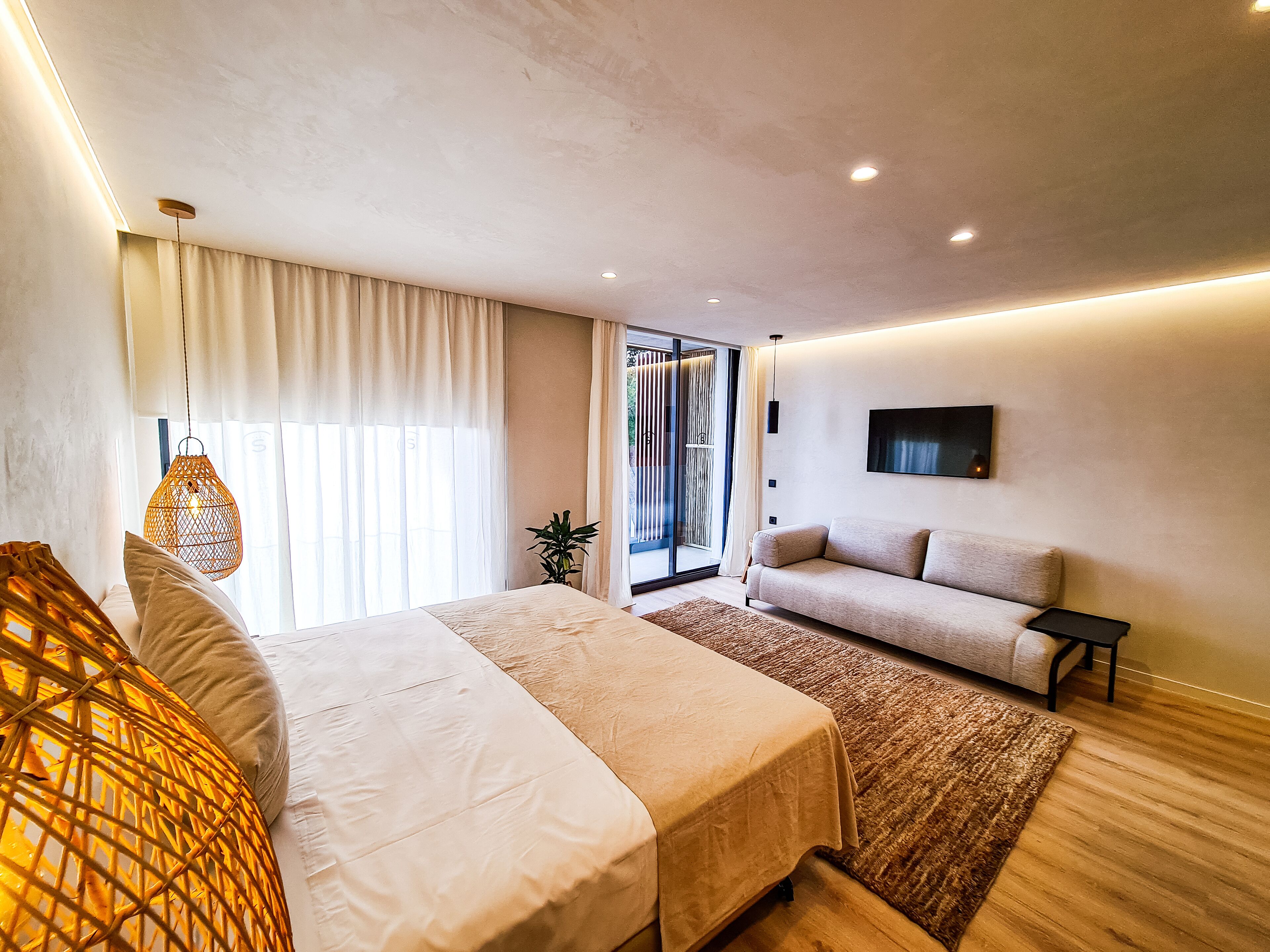 Un dormitorio moderno con cama, sofá, televisor y un ventanal con cortinas.