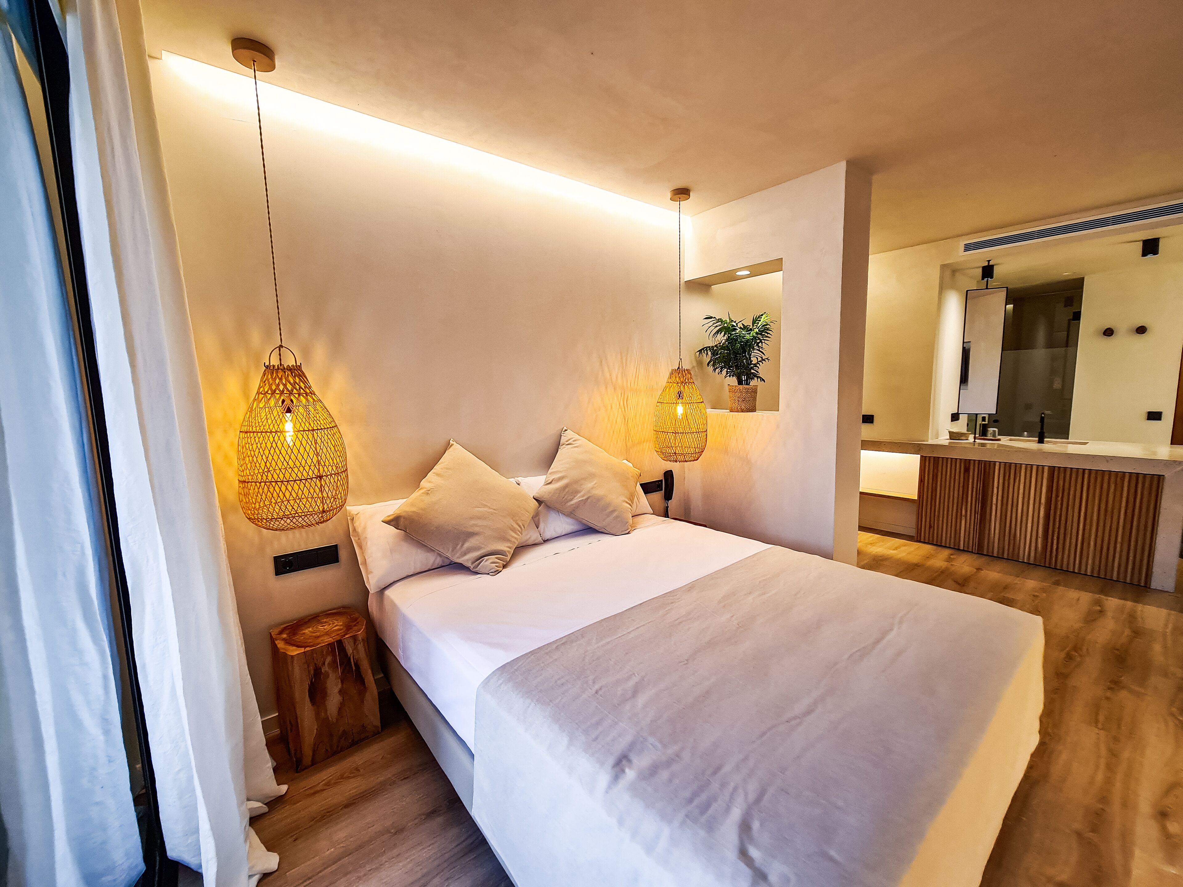 Un dormitorio moderno con una cama grande, lámparas de noche, un taburete de madera, un espejo y una planta.