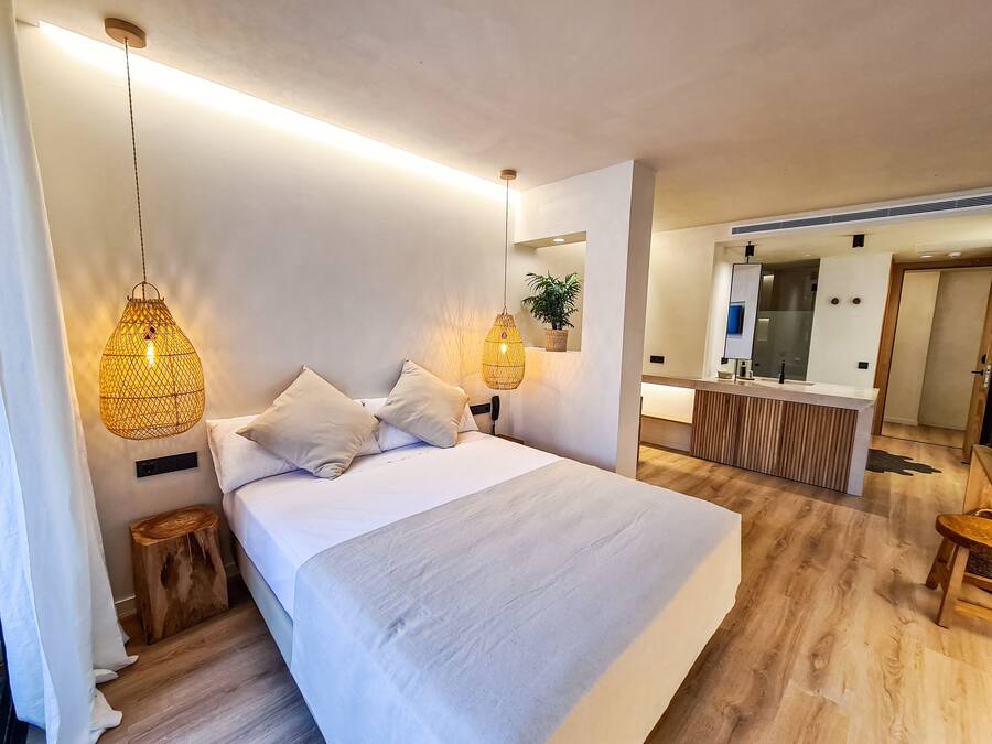 Una habitación de hotel moderna con una cama grande, dos lámparas de noche, un taburete de madera y una planta pequeña.