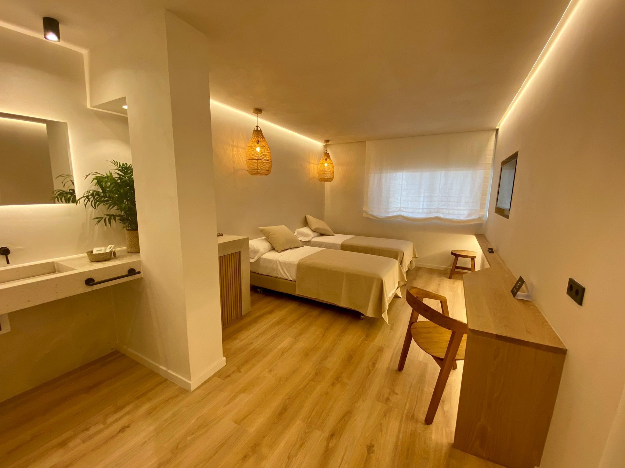 A modern bedroom with two beds, a wooden desk, a small table, a mirror, and a potted plant.