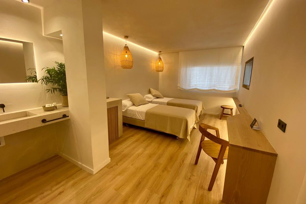 A modern bedroom with two beds, a wooden desk, a small table, a mirror, and a potted plant.