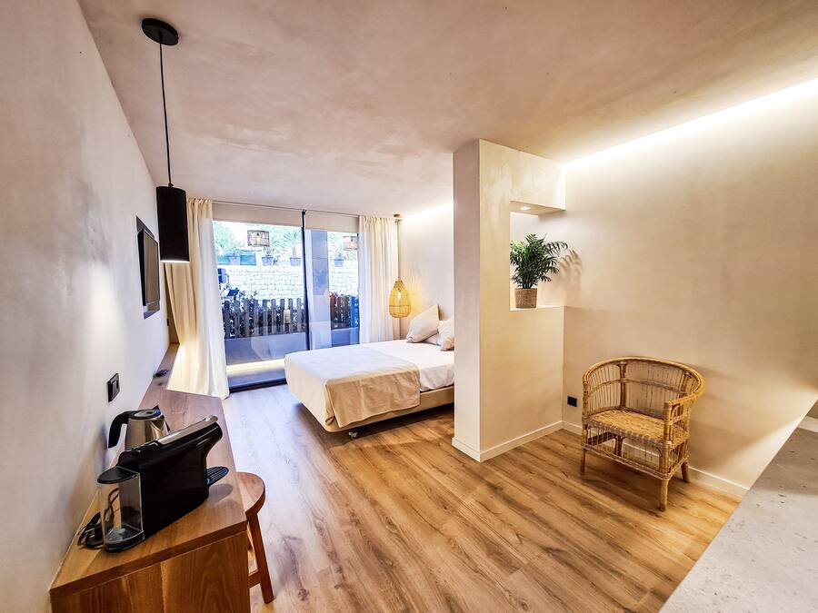 Habitación de hotel pequeña con cama, escritorio con hervidor, silla de mimbre y planta en maceta.