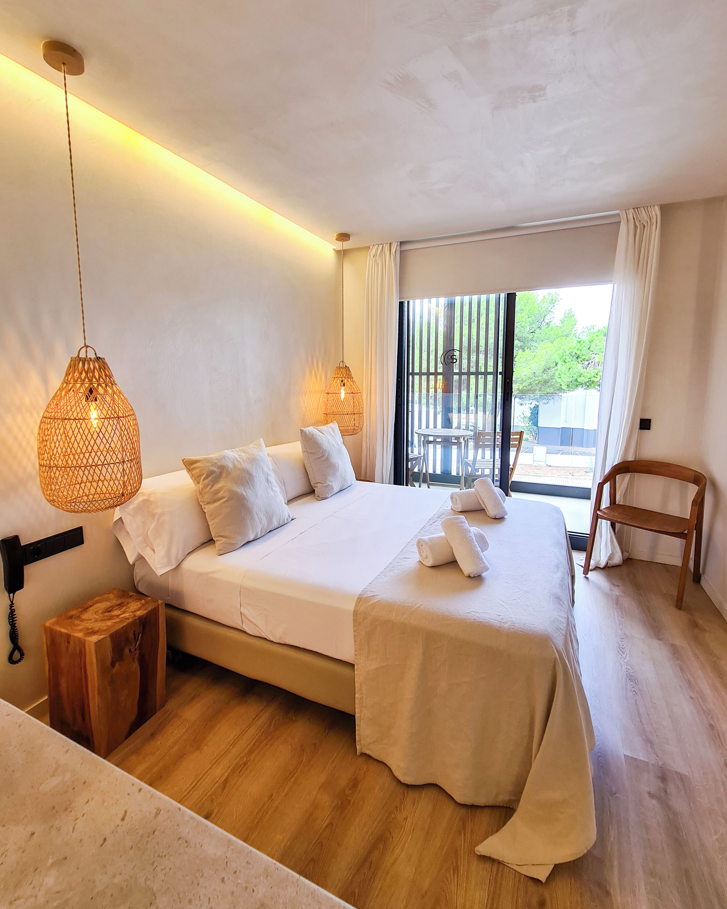 A bedroom with a bed, bedside lamps, a wooden stool, a chair, and a view through a window.