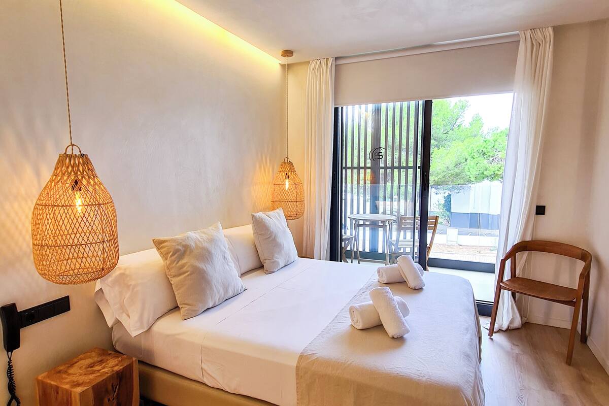 A bedroom with a bed, bedside lamps, a wooden stool, a chair, and a view through a window.