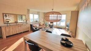 A modern living room with a wooden dining table, a large mirror, and a rattan pendant light. - Vacation apartment right by the sea near Callantsoog LekkerNaarZee (Callantsoog)