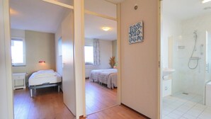 A bedroom with a bed, a bedside table, and a window. There is a bathroom with a shower and a sink. - Vacation apartment right by the sea near Callantsoog LekkerNaarZee (Callantsoog)