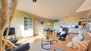 A modern living room with a sofa, coffee table, and TV. There is a kitchen area with cabinets and a dining table. - Vacation apartment right by the sea near Callantsoog LekkerNaarZee (Callantsoog)