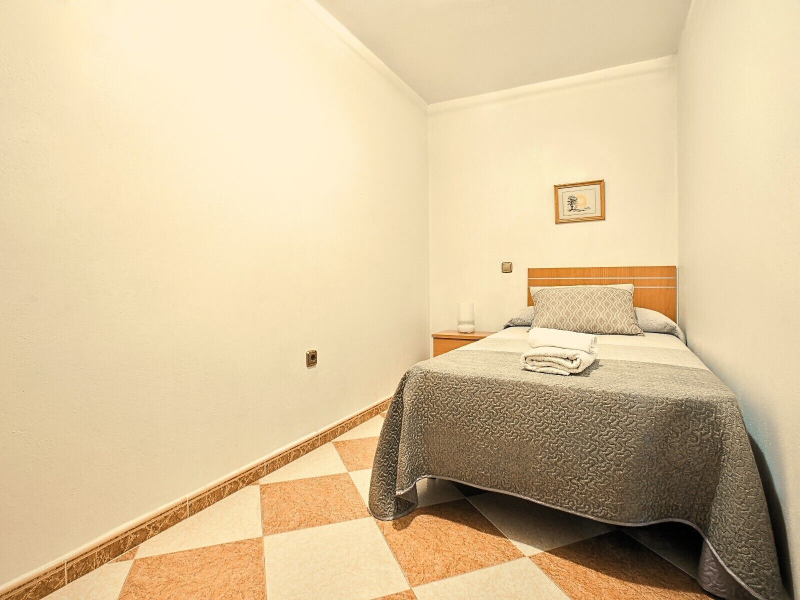 A small, neatly arranged bedroom with a bed, a nightstand, a framed picture on the wall, and a checkered floor.