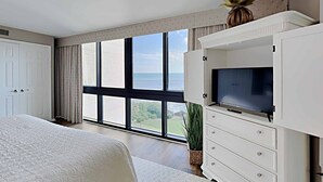 A bedroom with a large bed, a TV on a white cabinet, and a view of the sea through large windows. - Oceanfront Beach View, Large Pool, Updated! (Pawleys Island)