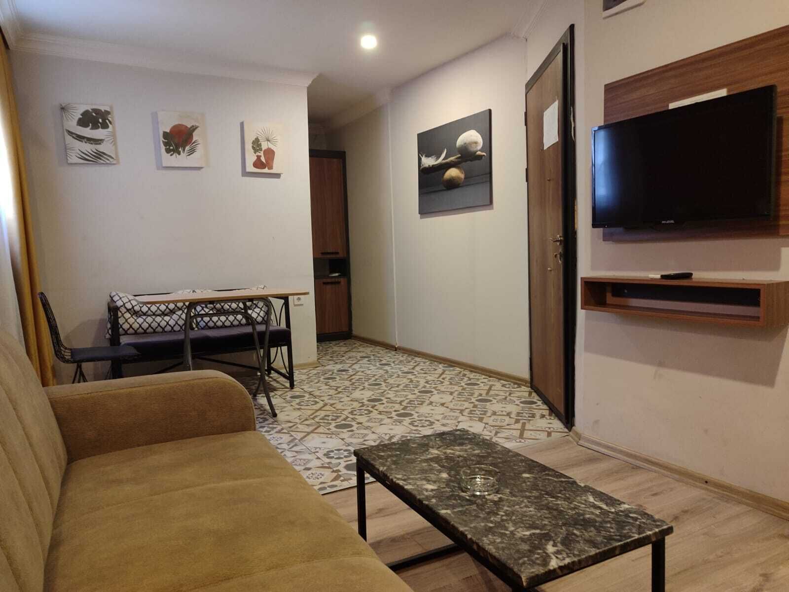 A living room with a sofa, a coffee table, a TV mounted on the wall, and a dining area with chairs and a table.
