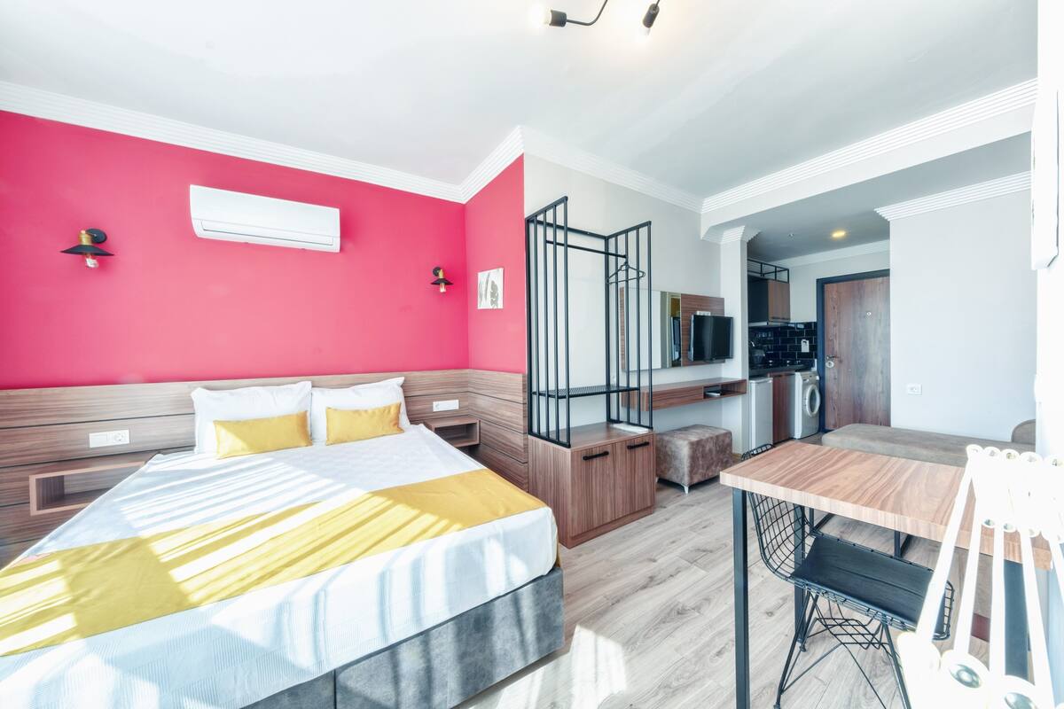 A modern hotel room with a large bed, a dining table, and a kitchenette.