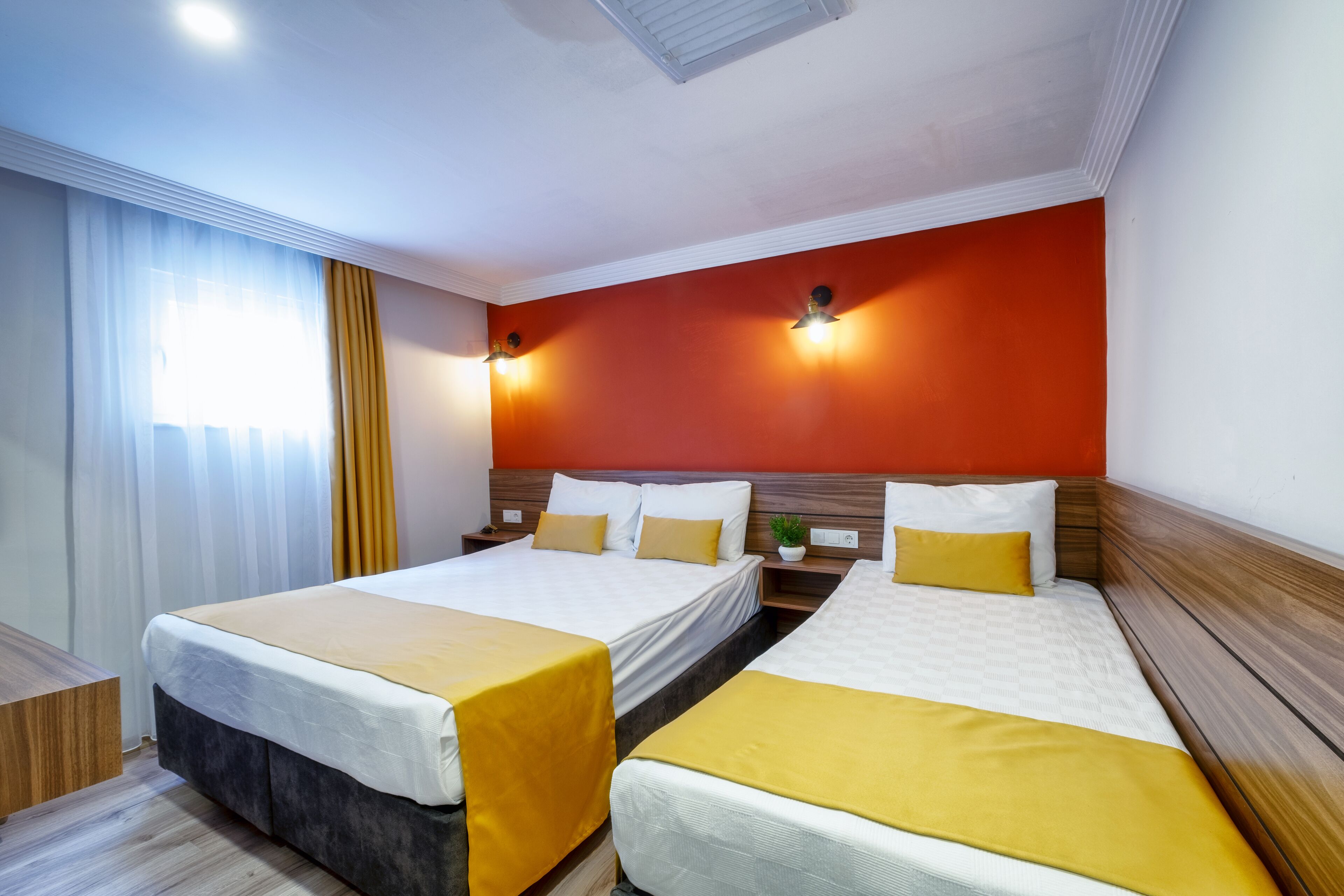 A hotel room with two beds, a wooden headboard, a large window with curtains, and wall-mounted lights.
