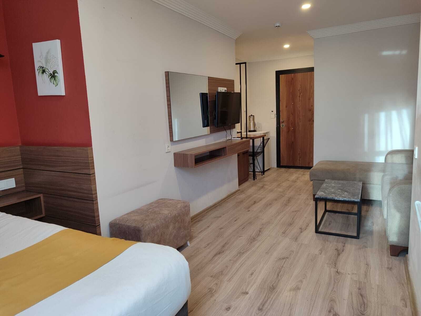 A hotel room with a bed, a sofa, a small table, a TV mounted on the wall, and a wooden door.