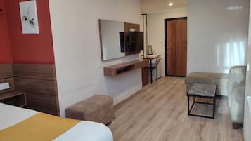 A hotel room with a bed, a sofa, a small table, a TV mounted on the wall, and a wooden door.