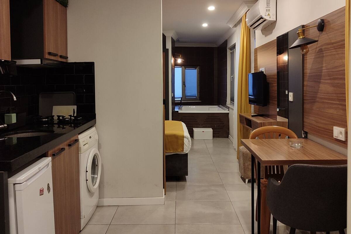 A modern kitchen with a washing machine, a dining area with a table and chairs, and a living space with a sofa and a TV.