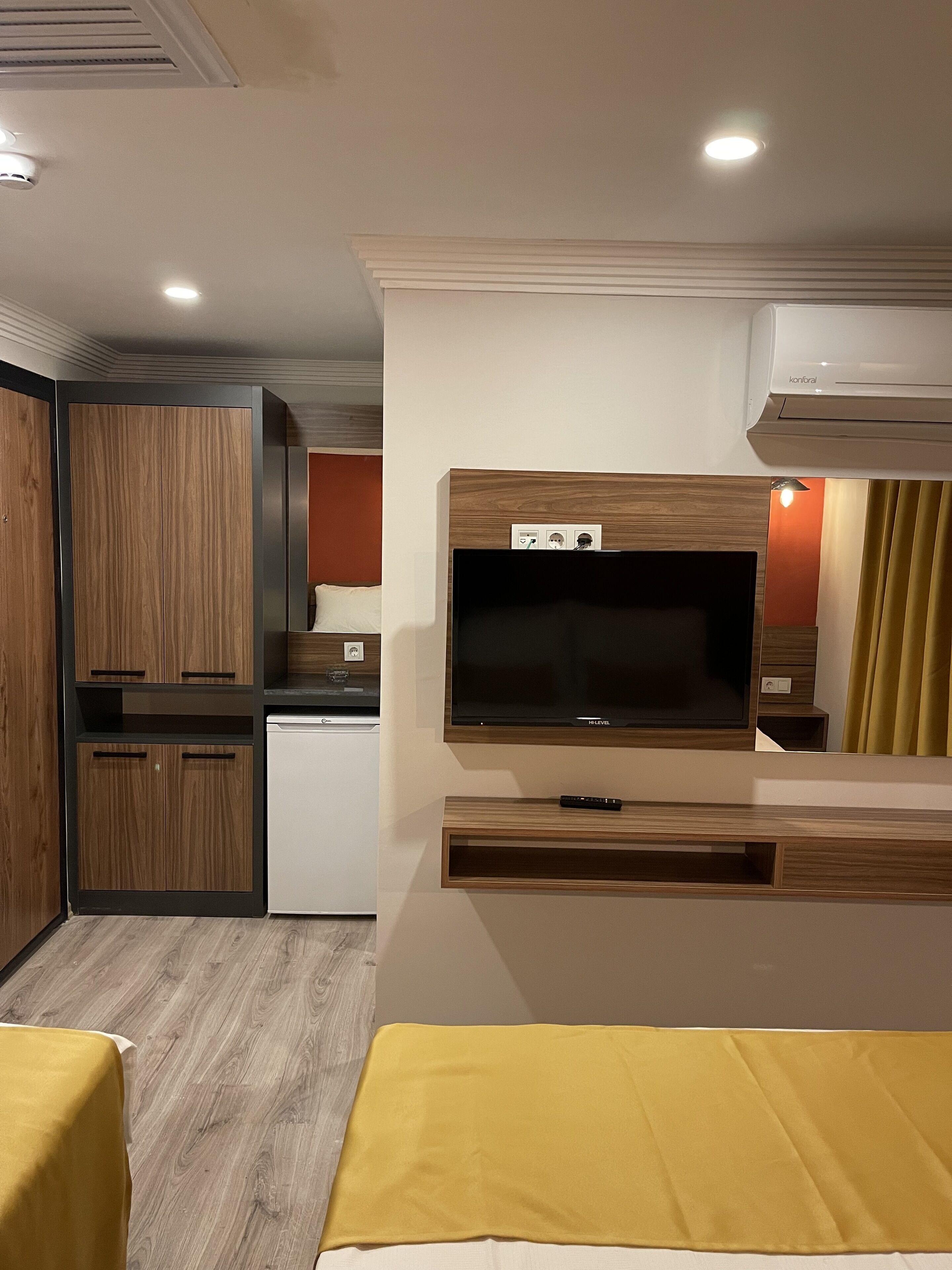 A modern hotel room with a wooden wardrobe, a built-in TV, a mini-fridge, and a yellow bedspread.