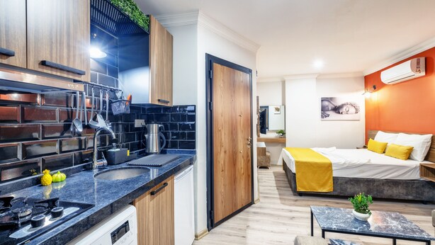 Una habitación de hotel compacta con kitchenette, una cama con ropa de cama amarilla, una pequeña mesa y una planta.