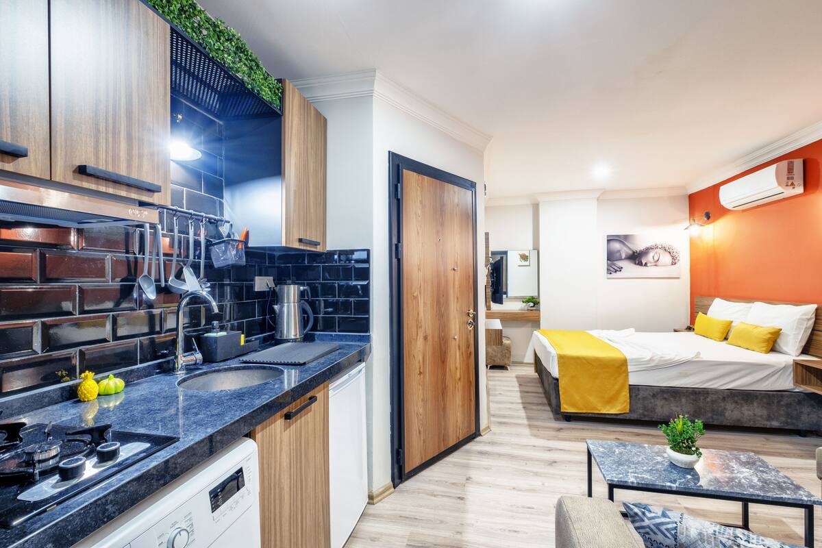 A compact hotel room with a kitchenette, a bed with yellow bedding, a small table, and a plant.