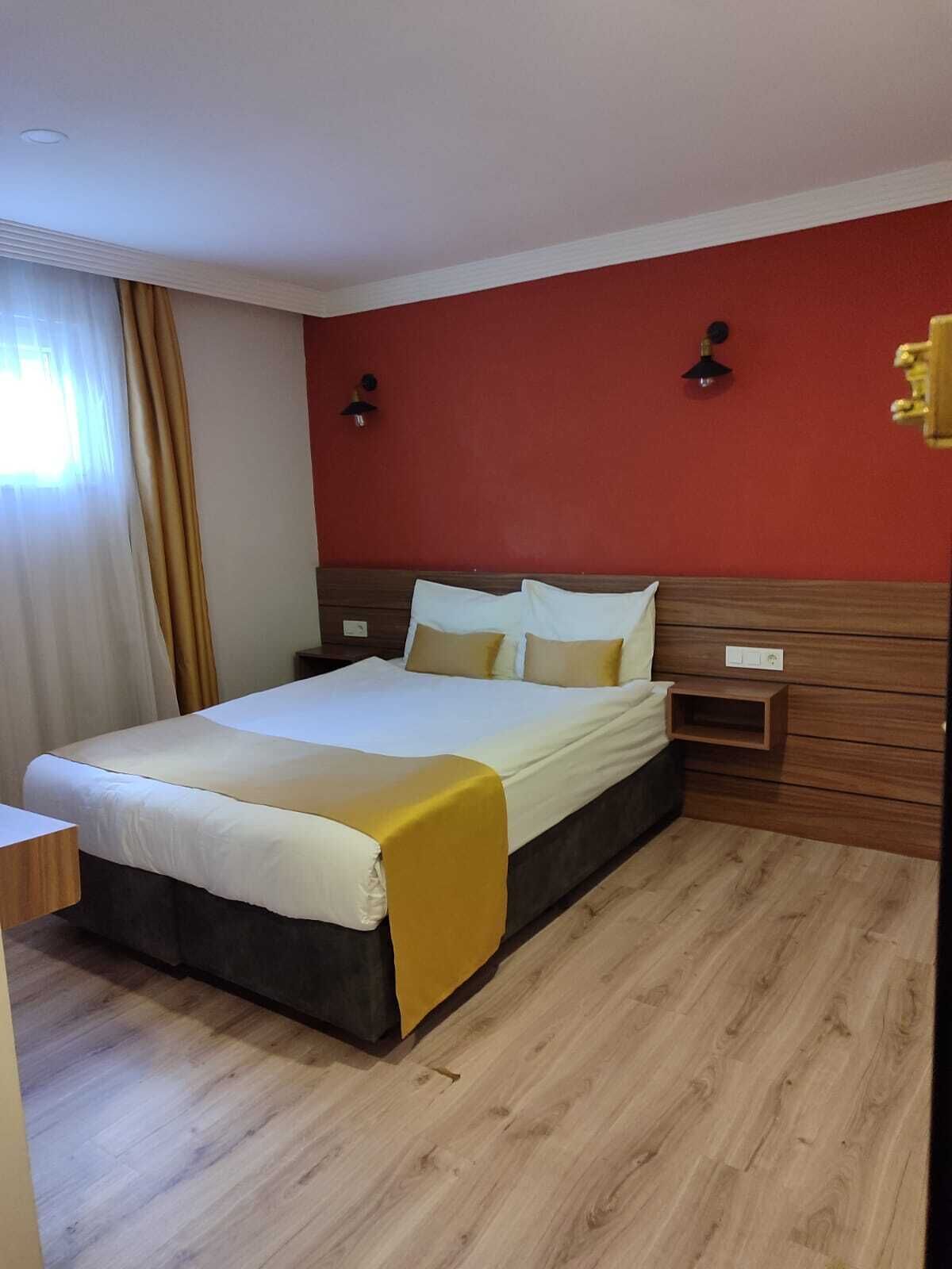 A hotel room with a bed, wooden floor, red walls, and two wall-mounted lights.