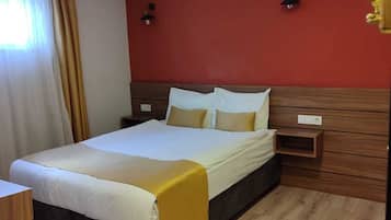 A hotel room with a bed, wooden floor, red walls, and two wall-mounted lights.