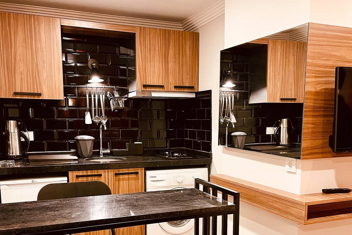 A modern kitchen with wooden cabinets, black tiled backsplash, stainless steel appliances, and a dining area with a metal chair and table.