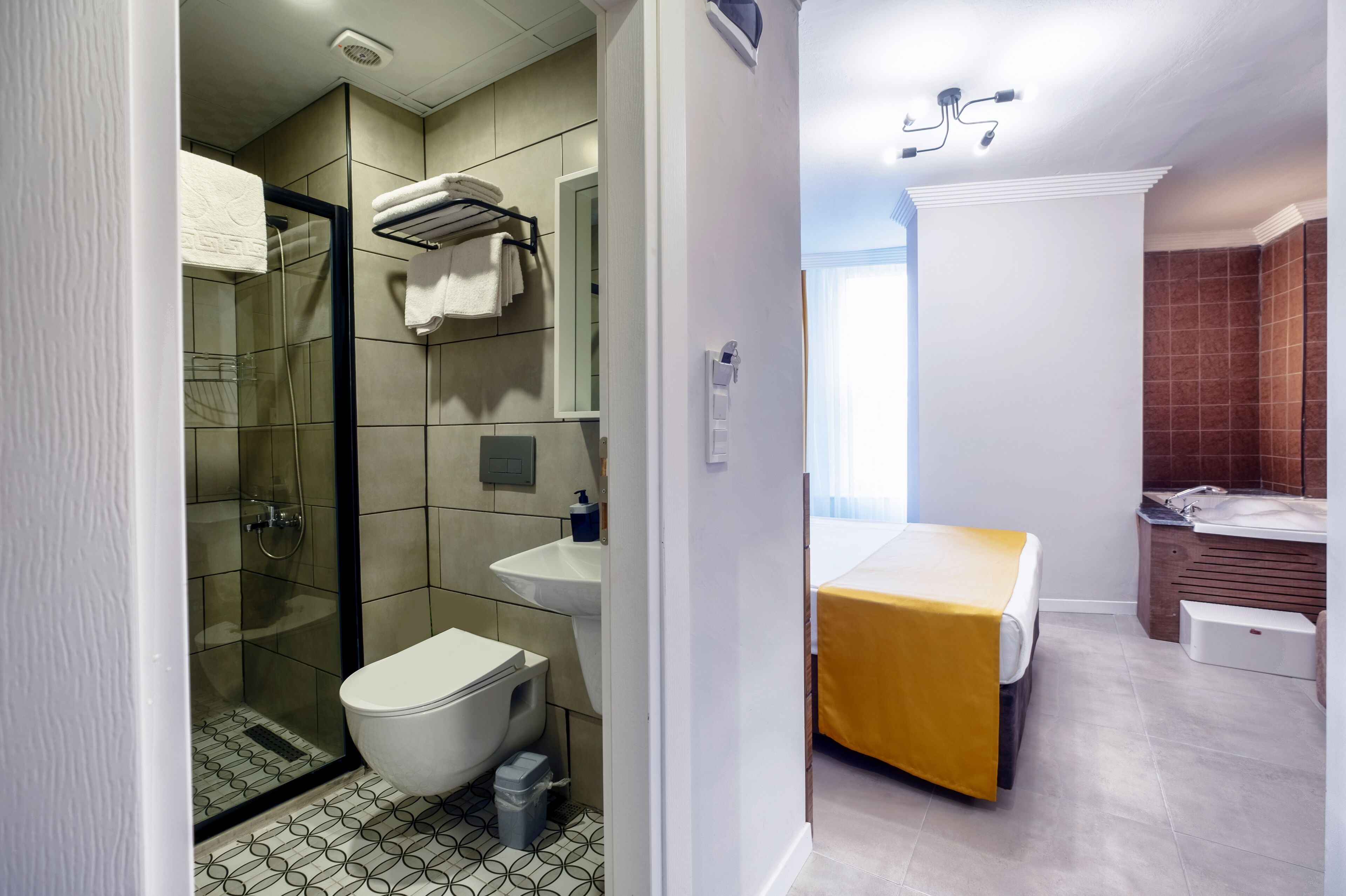 A modern bathroom with a glass-enclosed shower, a white toilet, a sink, and a bed with a yellow cover.