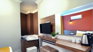 Habitación de hotel compacta con diseño moderno, que incluye un espejo grande, un televisor de pantalla plana, una pequeña zona de estar y una kitchenette con tetera y tazas.