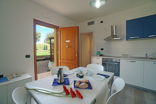 A compact kitchen with white cabinets, a stove, and a sink. There's a dining table with a white tablecloth, plates, and a coffee pot. A wooden door leads to an outdoor area. - Cascina al colle (Piombino)