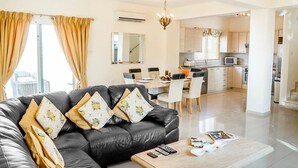 Een moderne woonkamer met een zwarte leren bank, een houten salontafel en een eethoek met een witte tafel en stoelen.
