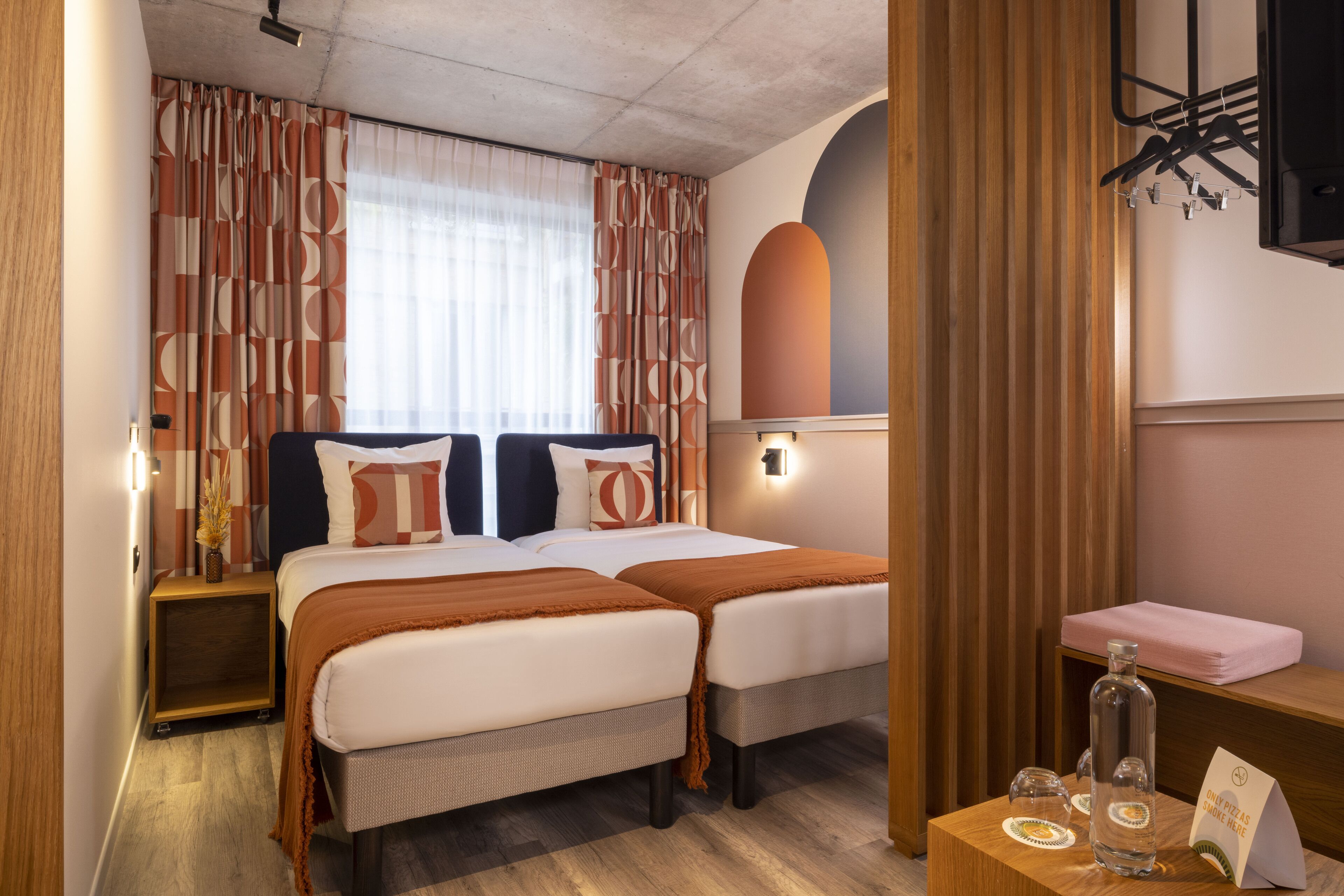 A hotel room with two beds, a wooden headboard, a small table with a bottle and a menu, and a mirror on the wall.