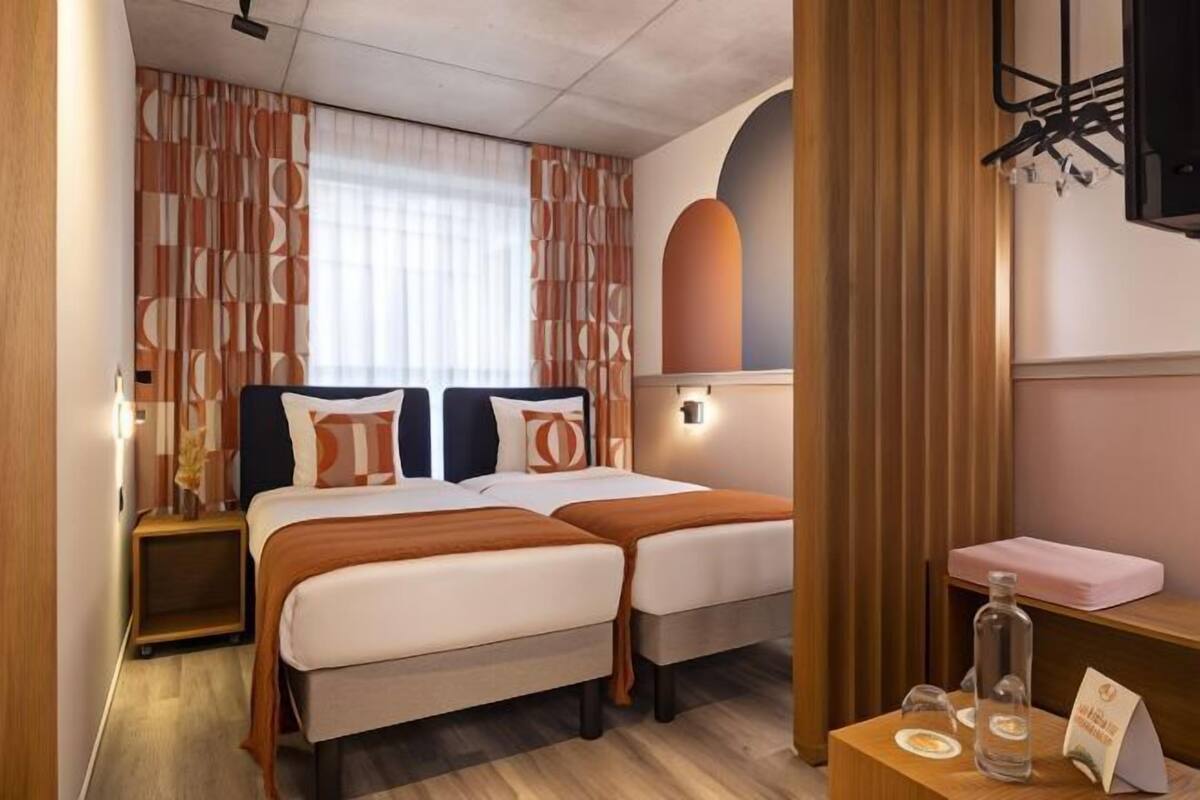A hotel room with two beds, a bedside table with a bottle and a menu, and a wall-mounted TV.