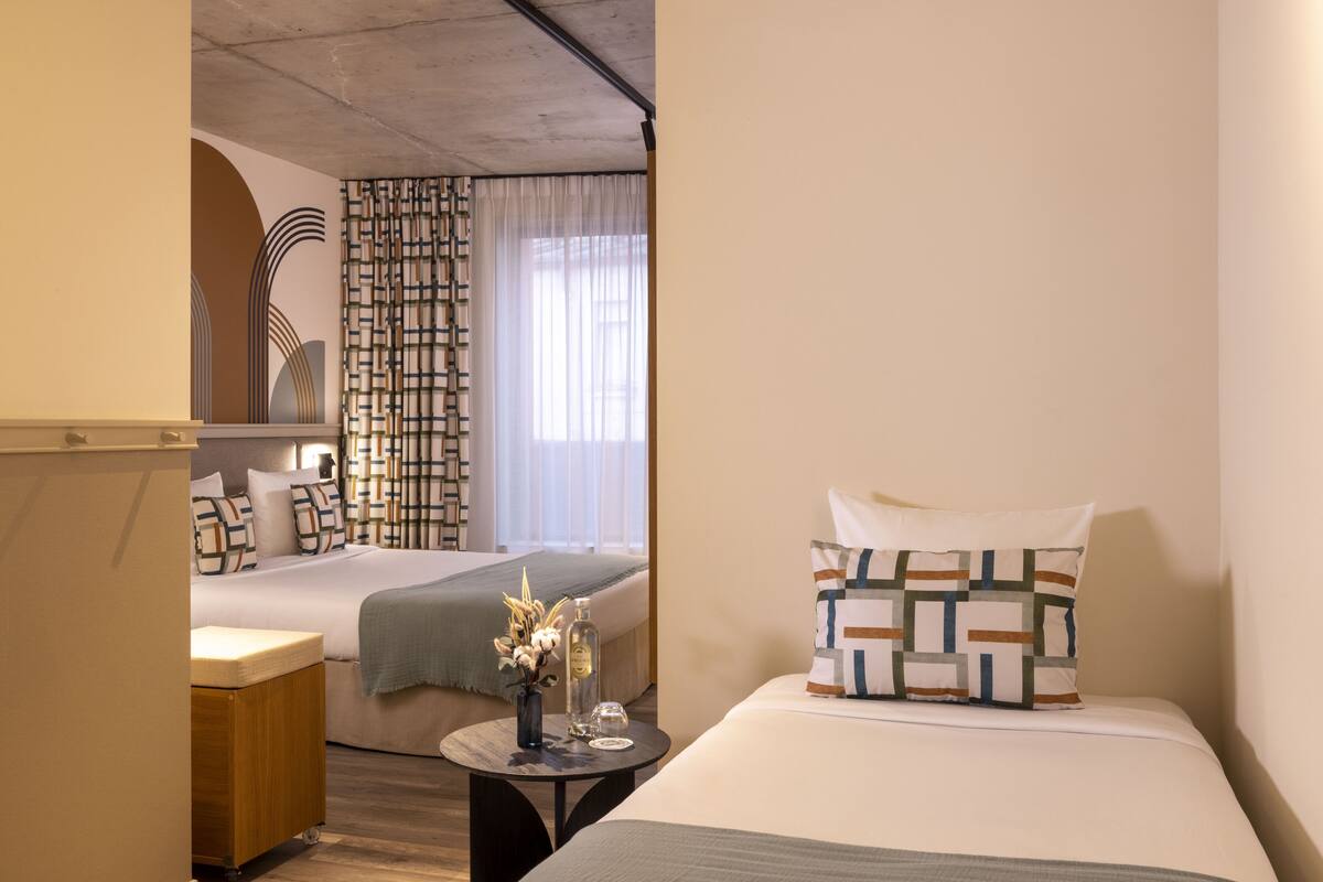 A hotel room with a bed, bedside table, and a view of another room with a bed and curtains.