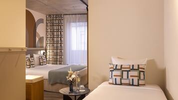A hotel room with a bed, bedside table, and a view of another room with a bed and curtains.